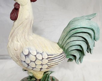 Pode incluir: Estatueta decorativa de galo em creme, bordô e verde. O galo tem um design detalhado e texturizado com uma crista e barbela bordô, uma cauda verde, e está empoleirado em uma base de pedra com folhagem verde.