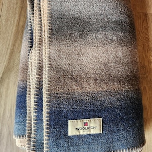 Puede incluir: Manta de lana doblada con un diseño degradado en tonos azules, grises y marrones. La manta tiene un borde cosido y una etiqueta rectangular que dice "WOOLRICH EST. 1830".