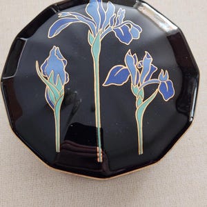 May include: Black octagonal trinket box with a gold rim. The lid features a design of three blue iris flowers with gold accents and green stems. The box has a glossy finish and a decorative floral theme.