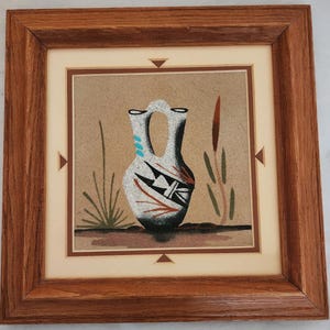 May include: A framed sand art piece depicting a white vase with black, red, and turquoise patterns on a tan backdrop. The artwork includes stylized plants and is set within a wooden frame with a cream-coloured mount.