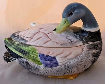 May include: A ceramic duck-shaped serving dish with a lid. The duck has a blue head, a green beak, and a pink and gray body. The wings are detailed with green, blue, and black feathers. The dish is approximately 10 inches long.