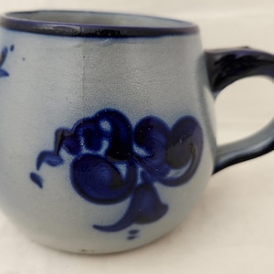 Puede incluir: Una taza de cerámica con base azul claro y borde y asa azul oscuro. La taza presenta diseños florales en azul oscuro. La taza tiene forma redondeada y un asa curva.