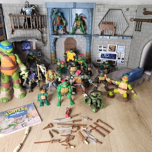 Può includere: Un set di gioco delle Tartarughe Ninja con un covo in pietra grigia a più livelli, numerose action figure di tartarughe verdi in varie pose, un personaggio di Shredder, un libro intitolato The First Battle e un assortimento di armi giocattolo.
