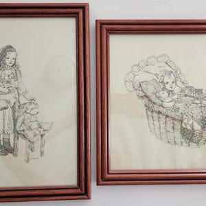 Könnte beinhalten: Zwei gerahmte Tuschezeichnungen. Der linke Rahmen zeigt ein Mädchen, das einen Teddybären hält und neben einem Teddybären auf einem kleinen Hocker steht. Der rechte Rahmen zeigt ein Baby in einem Korb.