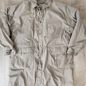 May include: A tan-colored, long coat with a button-up front, two large front pockets, and a collared neckline. The coat has long sleeves with buttoned cuffs and a drawstring at the collar. The coat is made of a lightweight material.