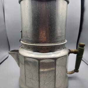 May include: A vintage silver-toned coffee percolator with a lid, spout, and a green-handled side handle. The percolator has a classic design with a cylindrical upper chamber and a faceted lower body. The metal has a worn, aged appearance.