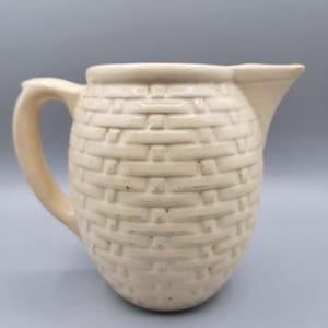 May include: Cream-coloured ceramic jug with a basket weave design. The jug has a handle and a spout. The overall shape is rounded, with a slightly flared top. The jug is likely vintage or antique.