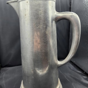 May include: A vintage, silver-colored pitcher with a handle and spout. The pitcher has a cylindrical body and a rounded base with three horizontal ridges. The handle is curved, and the spout is pointed. The pitcher appears to be made of metal.