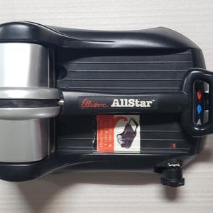 Puede incluir: Una máquina de troquelado manual Ellison AllStar negra y plateada con un mango horizontal. El dispositivo cuenta con una base negra con ranuras horizontales, un mecanismo de rodillo plateado y una etiqueta con texto rojo y blanco que dice Open here for Cutting Pad & Instructions.