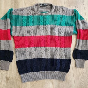 May include: A multi-colored sweater with a round neck and long sleeves. The sweater features horizontal stripes in gray, teal, red, and navy blue. The sleeves have a striped pattern in red, gray, and navy blue.