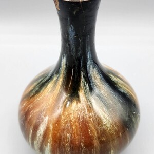 May include: A small, round ceramic vase with a narrow neck. The vase features a blend of colors, including brown, black, and cream, creating a marbled effect. The vase is approximately 6 inches tall.