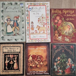 Op de afbeelding: Een verzameling vintage volkskunstboeken met kleurrijke illustraties. Titels zijn onder meer "Folk Art Expressions of Joy", "Holly Sprigs", "The Basics of Folk Art", "Chickens and Dumplin" en "A Folk Art Christmas."
