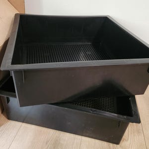 May include: Two rectangular black plastic trays stacked on top of each other. Each tray features a wide flat rim and a perforated grid base designed for drainage. The containers have solid vertical walls and a smooth finish on the exterior surfaces.