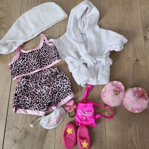 Puede incluir: Un conjunto de ropa de dormir y accesorios, que incluye una bata con capucha blanca, un conjunto de pijama con estampado de leopardo, zapatillas rosas con estrellas amarillas, una máscara para dormir y una pequeña bolsa rosa con el texto "Sweet Dreams".