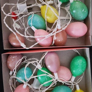 Peut inclure: Un ensemble de lumières de Noël en forme d'œuf de Pâques dans une boîte. Les lumières sont de différentes couleurs, notamment rose, vert, jaune et marron. Les lumières sont parsemées de points blancs.