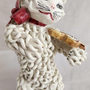 May include: White ceramic figurine of a cat playing a violin. The cat is covered in white, curly, textured fur. The cat has a red bow around its neck and is holding a brown violin.