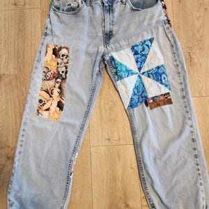May include: Light blue denim jeans with patches. One patch features a pinwheel design in shades of blue and white. Another patch displays a tattoo-style graphic of skulls and a woman. The jeans have a relaxed fit.