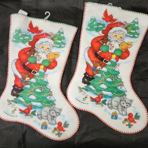 May include: Two white Christmas stockings with a Santa Claus design. Santa is wearing a red suit and holding a teddy bear and a rabbit. A green Christmas tree, birds, and a wolf are also featured. The stockings have a red trim.