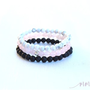 May include: Three stacked bracelets with black lava rock beads, pink rose quartz beads, and white howlite beads. The bracelets are on a white background.