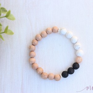 May include: A bracelet with light brown, white, and black beads. The bracelet has round, wooden-style beads, white marble-effect beads, and black lava beads. The bracelet is displayed on a white wooden surface.