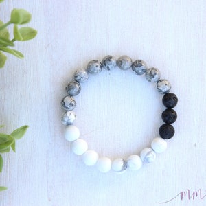 Puede incluir: Un brazalete de cuentas grises y blancas con cuentas de lava negras. El brazalete está hecho de piedras naturales y está diseñado para usarse en la muñeca.