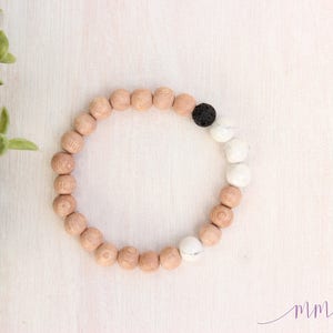 May include: A wooden beaded bracelet with a black lava bead and white beads. The bracelet is on a white wooden surface.