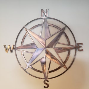 May include: A metal compass rose with a star design and the letters N, S, E, and W. The compass is a light brown color and has a rustic look.