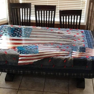 May include: A distressed American flag made of metal, with red, white, and blue stripes and stars, is displayed on a table covered in a blue and white patchwork fabric.