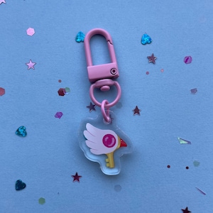 Peut inclure: Porte-clés rose avec un charme transparent représentant un oiseau rose avec une clé et une gemme rose. Le charme est attaché à un clip en métal rose.