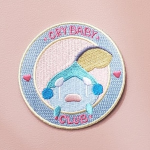 May include: A blue and yellow embroidered patch with a crying cartoon character. The patch is circular and has the text "CRY BABY CLUB" around the edge.