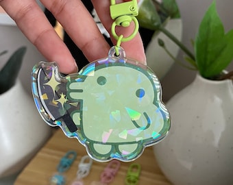 May include: A light green, holographic acrylic keychain featuring a cartoon character holding a knife. The keychain has a lime green clasp and ring. Additional keychains are visible in the background.