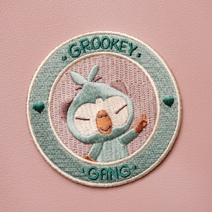 Puede incluir: Un parche bordado verde y blanco con un mono de dibujos animados verde y marrón haciendo un signo de paz. El parche dice "Grookey Gang" en un círculo.