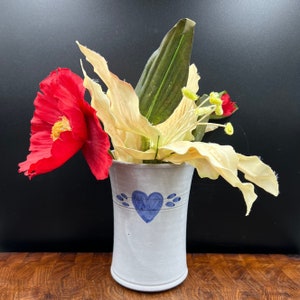 May include: A white ceramic vase with a blue heart design, holding a bouquet of red and white flowers. The vase has a textured surface and a slightly flared opening.