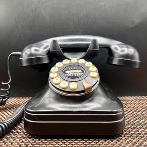 May include: A black desk telephone featuring a curved handset resting on a base with a circular push-button keypad. The keypad includes light-colored buttons with numbers and letters, and the center dial displays the text FLASH and REDIAL.