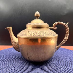 May include: A vintage brass teapot with a decorative floral pattern on the lid and spout. The teapot has a curved handle and a small finial on top. The brass has a warm, golden hue, and the teapot sits on a blue mat.