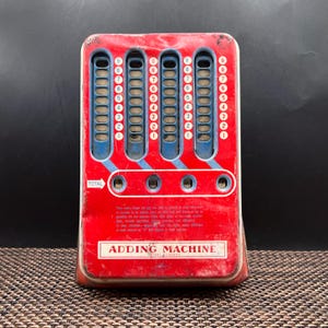 May include: A vintage red metal adding machine featuring four vertical blue sliding columns with numbers one through nine. The device includes a total display window, instructional text, and the words ADDING MACHINE printed in white on the front surface.