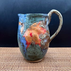 May include: A ceramic pitcher featuring a prominent embossed maple leaf design in shades of orange and red. The body of the vessel displays a mottled glaze with blended tones of blue, green, and brown, complemented by a curved, textured handle.