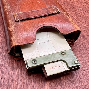 May include: A vintage slide rule partially extended from a worn, brown leather case. The slide rule has white and black markings, with a metal frame and <4092> text. The case shows signs of age and use.