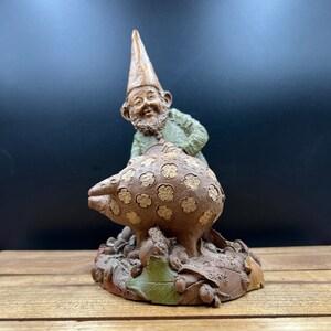 May include: A whimsical garden gnome figurine. The gnome, with a pointed hat and green jacket, is positioned behind a brown, mushroom-shaped object adorned with floral patterns. The base features leaves and acorns, creating a charming, decorative piece.