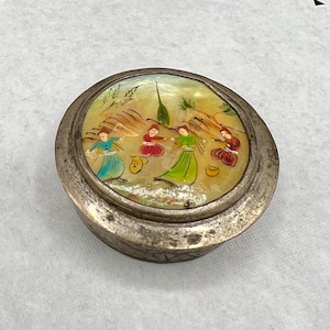 May include: A round, silver-toned trinket box with a painted lid. The lid features a scene of figures in colorful clothing dancing in a landscape with yellow and green hues. The box has a tarnished appearance.