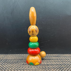 May include: Wooden stacking toy in the shape of a rabbit. The rabbit has a long, oval head and a round face with painted eyes and a nose. The body is made of colorful, round pieces: yellow, green, and red. A small wooden ball sits next to the rabbit.