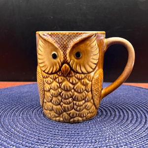 May include: A brown ceramic mug shaped like an owl, featuring a textured surface that mimics feathers, two prominent round eyes, a pointed beak, and a curved handle on the right side. The mug has a glossy finish with darker brown shading.