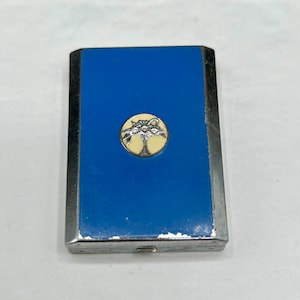 May include: A rectangular, vintage cigarette case with a bright blue enamel top and silver-toned metal trim. A circular emblem with a tree design is centered on the blue surface. The case has a small push-button clasp.