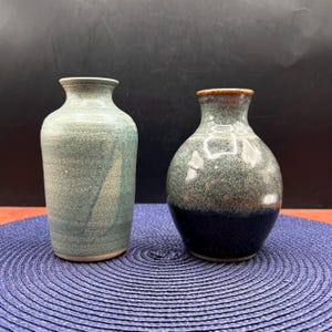 May include: Two ceramic vases with distinct shapes and finishes. The left vase is tall and cylindrical with a matte, speckled teal glaze. The right vase is bulbous and rounded, featuring a glossy, mottled green top and a solid dark blue base.