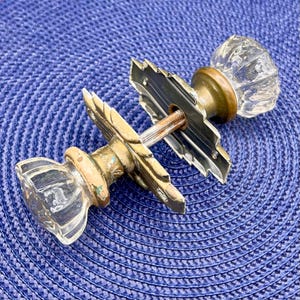 May include: Two antique glass door knobs with brass and metal hardware. The knobs are clear, faceted, and have a vintage aesthetic. The backplates are ornate with a decorative design. The hardware is aged with a patina.