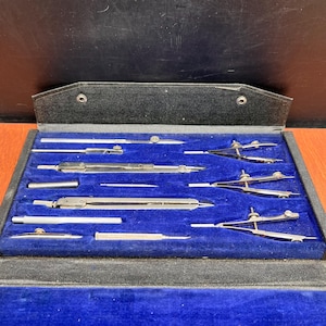May include: A set of silver-toned metal drafting instruments arranged in a black case with a blue velvet interior. The collection includes various compasses, dividers, and extension bars with adjustable screws and pointed metal tips for precision.