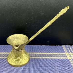 May include: A brass coffee pot with a long, ornate handle. The pot has a wide, open top and a rounded base with floral patterns. The handle is also decorated with etched designs. The pot is likely used for making Turkish or Greek coffee.