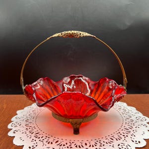 May include: A ruby red glass basket with a ruffled edge and a decorative gold-toned metal handle. The basket sits on a gold-toned metal base with three feet. The handle has an ornate filigree design.