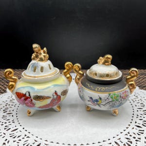 May include: Two ornate, small ceramic incense burners. One is white and yellow with a painted scene of figures, the other is black and white with a bird and floral design. Both have gold accents and lids.