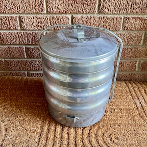 May include: A cylindrical, multi-tiered aluminum food carrier featuring a stackable design with three distinct compartments. It includes a secure, fitted lid with a central metal handle and a sturdy, hinged wire bail handle for easy transport.